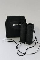 Photo of small Tasco binoculars positioned in front of black carrying pouch with Tasco logo, showing binoculars with rubberized textured grips and an attached neck strap.