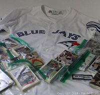 Photo showing white Blue Jays Majestic baseball jersey with visible front logos and some small stains, surrounded by several plastic baggies containing sports cards.