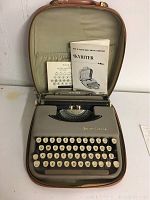 Vintage Smith Corona Skyriter portable typewriter in gray metal with white keys, inside a brown lightweight travel case, with instruction manuals placed behind the typewriter.