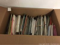 Top view of a large cardboard box filled with assorted stationery and greeting cards, showing different colors and sizes, some bundled with rubber bands.