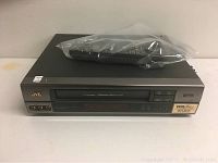 Black JVC VCR player with remote control in plastic bag on top