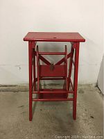 Front view of red wooden stool showing folded stool form with a rectangular seat and cross braces.