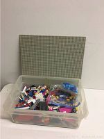 Clear plastic bin filled with large grey Lego base plate standing behind it.