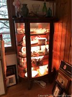 The full curio cabinet with internal lighting on, showing various glass and ceramic items inside and on top.