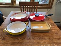 Overall view of 5 enamelware cookware pieces on wooden table including red and yellow pans with lids, and beige baking dish.