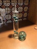 Tall glass cylinder thermometer containing liquid with five floating greenish-blue glass bulbs with metal tags and accompanying greenish blown glass ball.