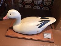 Full view of the carved white duck decoy with black wing markings mounted on a polished wooden base beside a conservation stamp plaque.