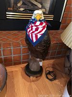 Full view of bald eagle sculpture standing on wooden floor in front of brick hearth; eagle wears American flag bandana.