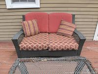 Front view of the outdoor loveseat showing red back cushions, patterned seat cushions, striped throw pillows, and dark woven wicker frame. Part of the wicker table is visible in the foreground.