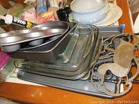 Stacked metal and glass baking sheets to include muffin pan, cookie sheets, and other bakeware along with a metal trivet and a large ceramic tureen with lid on top of a plate.