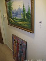 Photo showing framed landscape painting hung on wall, smaller print of three women leaning against wall below it.