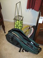 Tennis rackets stacked inside a green and black Head tennis bag alongside a metal ball hopper filled with tennis balls