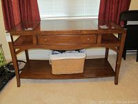 Front view of medium brown wood console table with glass top panels, central drawer with brass knobs, and bottom shelf. Carpet floor and window with curtains in background.