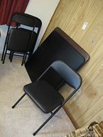 Black folding card table leaning against wood paneled wall, with one black folding chair positioned in front and three chairs stacked nearby.