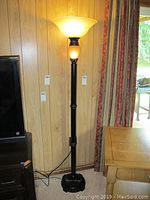 Full view of the 68 inch tall black metal floor lamp with frosted glass shade lit up, positioned beside wooden panel wall and curtain.