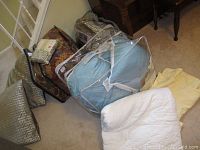 Lot showing packaged comforter sets, pillows on the floor next to wooden furniture.