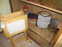 Photo showing three wooden TV trays with white tile tops and folding stand, and adjacent shelf with various serving bowls and plates including a black metal serving stand with white plates.