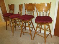 Four oak bar stools with carved wooden backrests and burgundy seat cushions, arranged side by side against a wall.