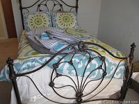 Full view of the metal bed frame's footboard with bedding and pillows, showing the bedspread's colorful circular and leafy patterns in green, blue, and white.