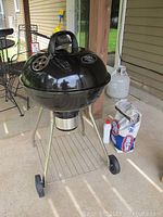 Full view of black Masterbuilt Pro charcoal grill on metal frame with wheels, including charcoal briquettes bag, bottle of fluid, and grill brush on the ground.