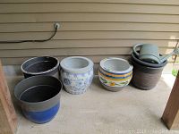 Set of 5 planters including 2 ceramic and 3 vinyl, with watering can on top of ribbed vinyl planter. All placed on concrete floor beside house siding.