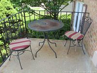 Round metal mesh table with two matching metal scroll-back chairs with striped cushions on a concrete patio.