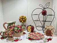 Full set of apple-themed ceramic teapot, creamer, sugar bowl, plate, candle holder, artificial apple tree, and black metal apple-shaped cup rack arranged on a white surface with apple motif cloth beneath.