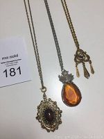 Three pendant necklaces laid out: golden filigree with brown stone, teardrop amber-orange stone with silver floral cap, silver-tone flower with gold drops