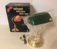 Photo showing the solid brass desk lamp with green shade wrapped in plastic next to its original black Universal branded box.