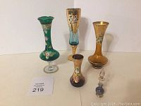 Four vintage Italian Murano glass vases and cordial glasses with gold accents and hand-painted floral designs, positioned side by side on a white surface.