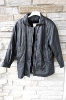 Front view of XL black leather jacket with zipper and snap-button closure, pockets, and elastic cuffs, hanging against stone wall.