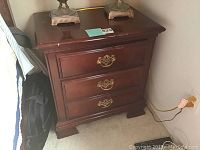Photo showing one of the two wooden night stands with three drawers and ornate metal handles, placed next to a bed and wall outlet, with visible scratches and wear.