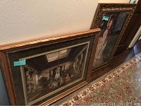 Photo showing two large framed art pieces leaning against a wall on a patterned rug, one landscape with an indoor scene and another portrait with a woman in a dress.