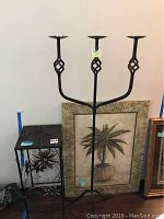 Photo of black iron candle stand with three arms, metal table with flower design and woven top, and framed palm tree art behind them.