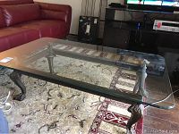 Glass top coffee table showing clear glass rectangular surface and detailed claw foot iron base on a carpeted living room floor beside a red couch.