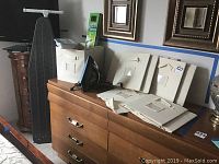 View of eight beige fabric storage bins, black Sunbeam iron, ironing board leaning against wall, all arranged on and around a wooden dresser.