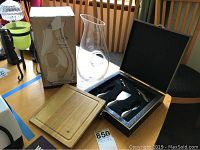 Photo showing the pierced glass pitcher with original box on the left and the opened black hinged box containing cheese utensils on the right on a wood table