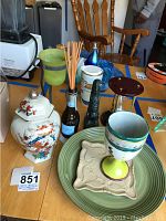 Overview of various ceramic, glass, and figurine items arranged on a table showing a floral ceramic jar, ceramic goblet, drinking cups, serving plate, diffusers, and figurine