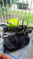 Military backpack, Motorcycle Tour Master bag, and High Sierra fanny pack on ground next to green buckets and metal railing.
