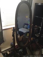 Frontal view of oval floor mirror with wooden frame and stand in a room with natural light from window.