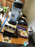 White Vitamix 5000 blender on wooden table with clear blending container, black lid, and tamper tool. Whole Food Recipes cookbook included.