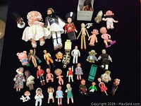 Photo showing the entire lot of approximately 39 miniature vintage plastic dolls and figures arranged on a black cloth surface, including soldiers, babies, cartoon characters, and assorted dolls in various clothing and poses.