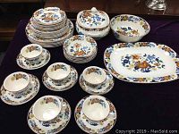 Photo showing full set of plates, cups, saucers, covered tureen, platter, and vegetable bowl arranged on table with Kismet floral pattern primarily in brown, blue, yellow, and green.