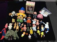 Full view of assortment of vintage dolls and toys spread out on black cloth. Includes Spiderman doll, army outfit, small dolls, and boxed doll.