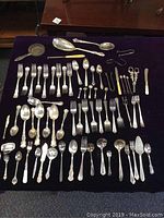 Overview of the entire mixed lot of vintage silver plate cutlery laid out on a dark cloth showing forks, spoons, dessert spoons, serving pieces and specialty utensils in two different decorative patterns.