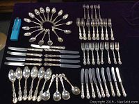 Full set of the cutlery laid out on dark fabric background, showing the variety of teaspoons, knives, dessert spoons, pickle forks, forks, cake forks, and butter knives in Regency pattern handles.
