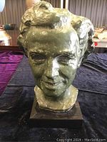 Front view of greenish bronze bust sculpture showing detailed facial features and hair, mounted on dark square base.