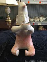 Front view showing seated humanoid clay figure with bowl held in hands, painted in pink and white.
