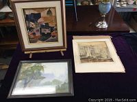 Three watercolours displayed on a table, two framed and one unframed on paper, with various surrounding furniture
