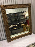 Full view of the antique framed mirror leaning against a wall, showing the entire reflective glass and the ornate gold frame in a room with shelving in background.